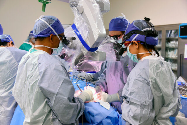 UF Health oral and maxillofacial surgeons in blue scrubs peer into a operating microscope during surgery.
