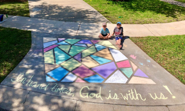 Two children sit with chalk art of a stained-glass window with the words, In hard times, God is with us!