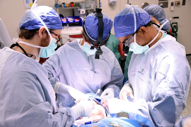 Three UF Health surgeons in blue scrubs operate on a patient.